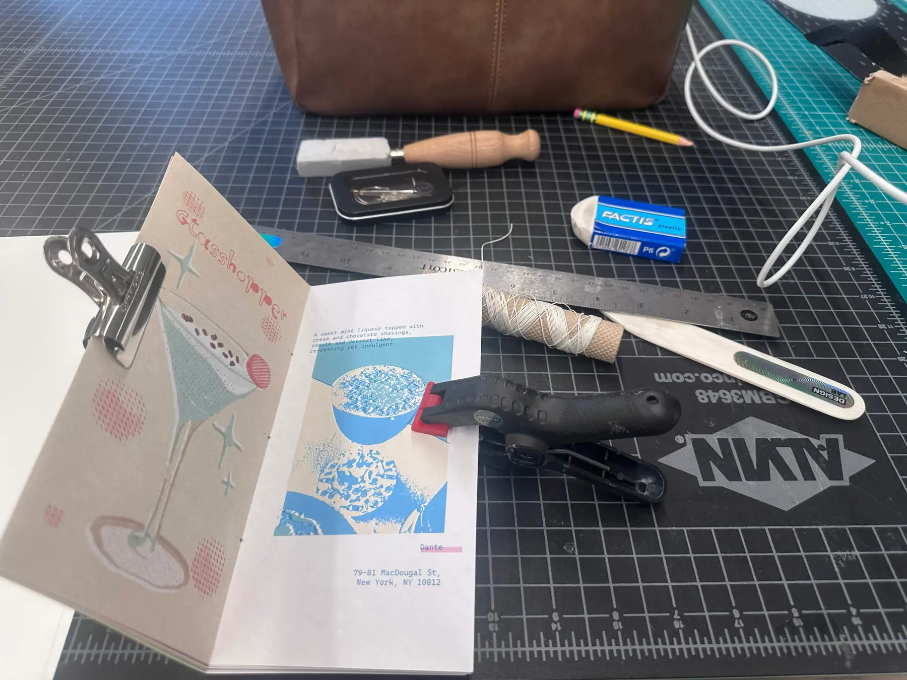 A printed copy of the zine open on a cutting mat, clipped at the spine, with an awl, bone folder, waxed thread and a steel ruler laid out beside it
